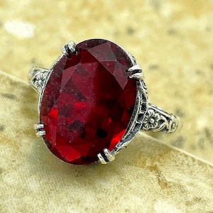 May include: A silver ring featuring a large, oval-cut, deep red faceted gemstone held by four prongs. The metal band displays an intricate, raised floral pattern along its sides, providing a textured appearance to the silver setting.