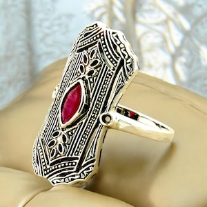 May include: A silver ring with an intricate design featuring a large, faceted, ruby-red gemstone. The ring has a wide band and a rectangular shape.