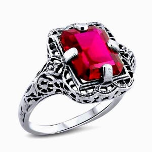 May include: A silver ring with a large, rectangular, bright red gemstone. The ring has an ornate, filigree design around the gemstone and on the band. The ring is set against a white background.