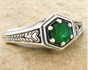 May include: A silver ring featuring a round, vibrant green gemstone set within a hexagonal frame. The band displays intricate, engraved patterns including chevron and heart-like motifs along the sides, with a polished metallic finish throughout.