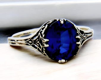May include: A silver-toned ring featuring a large, oval-cut deep blue gemstone set in a decorative, filigree-style metal band. The metalwork surrounding the central stone has a dark, oxidized finish that highlights the intricate, textured patterns.