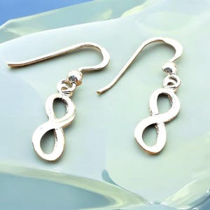 May include: A pair of silver infinity symbol earrings with a hook closure. The earrings are on a white surface.