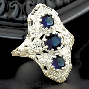May include: A silver-toned ring featuring an elongated, openwork filigree design. The ring is set with three round, deep blue stones arranged vertically, accented by smaller, clear, round stones embedded within the intricate metalwork.