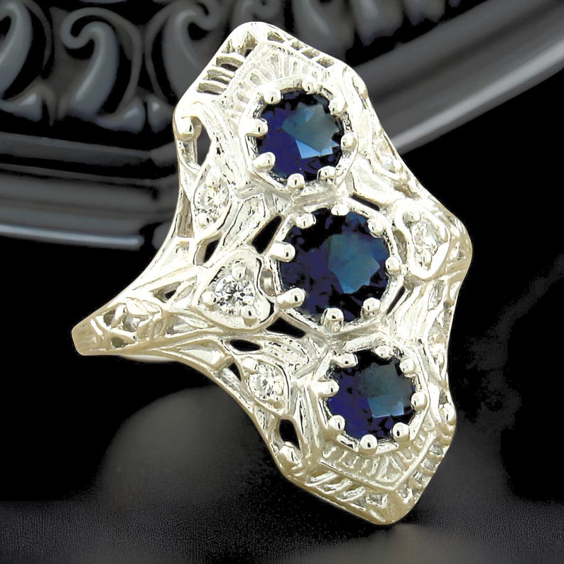 May include: A silver-toned ring featuring an elongated, openwork filigree design. The ring is set with three round, deep blue stones arranged vertically, accented by smaller, clear, round stones embedded within the intricate metalwork.