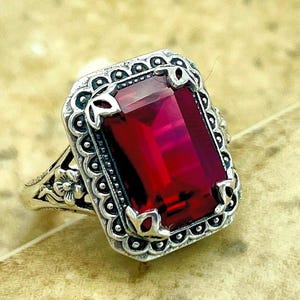 May include: A silver-toned ring featuring a large, rectangular, deep red gemstone set in a decorative, scalloped metal frame. The band displays intricate floral engravings on the sides, with a polished finish that highlights the detailed metalwork.