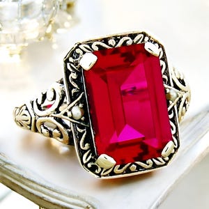 May include: A vintage-style ring with a large, bright red emerald-cut gemstone set in a gold filigree band. The band is adorned with two small pearl accents.