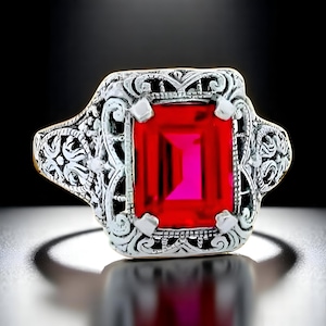 May include: A silver ring with an ornate filigree design. The ring features a large, rectangular, bright red gemstone. The gemstone is the focal point, set in a detailed, vintage-style setting. The ring is displayed against a dark background.