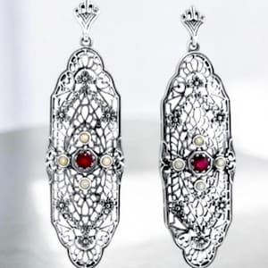 May include: A pair of long, silver-toned drop earrings featuring an intricate, openwork filigree design. Each earring is centered with a single, round, deep red stone surrounded by four small, round, white pearls in a cross-like arrangement.