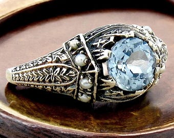 May include: A silver ring with a large blue gemstone surrounded by smaller white pearls. The ring has an intricate design with floral motifs.