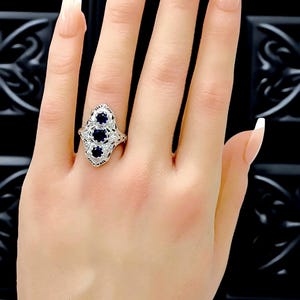 May include: A silver-toned ring featuring three round, deep blue sapphire stones arranged vertically in a marquise-shaped setting with intricate metal filigree detailing and small clear accent stones embedded along the band's decorative frame.