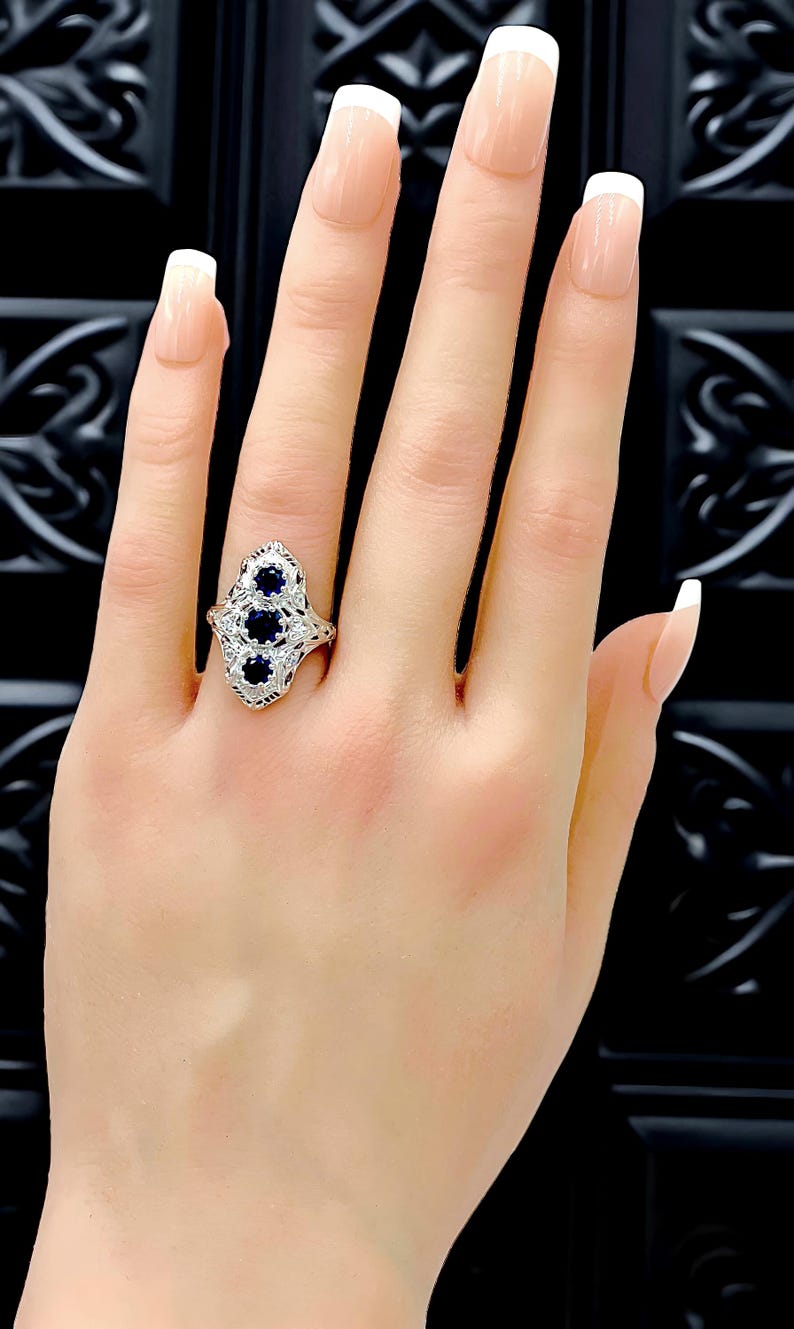 May include: A silver-toned ring featuring three round, deep blue sapphire stones arranged vertically in a marquise-shaped setting with intricate metal filigree detailing and small clear accent stones embedded along the band's decorative frame.