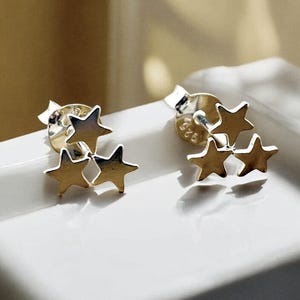 May include: A pair of gold-colored star-shaped stud earrings. Each earring features a cluster of three stars, with a post and backing for secure wear. The earrings are displayed on a white surface.
