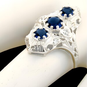 May include: A silver-toned ring featuring three round, deep blue faceted stones set in a row. The metal band displays an intricate, openwork filigree design with small, clear, colourless accent stones embedded throughout the decorative metal frame.