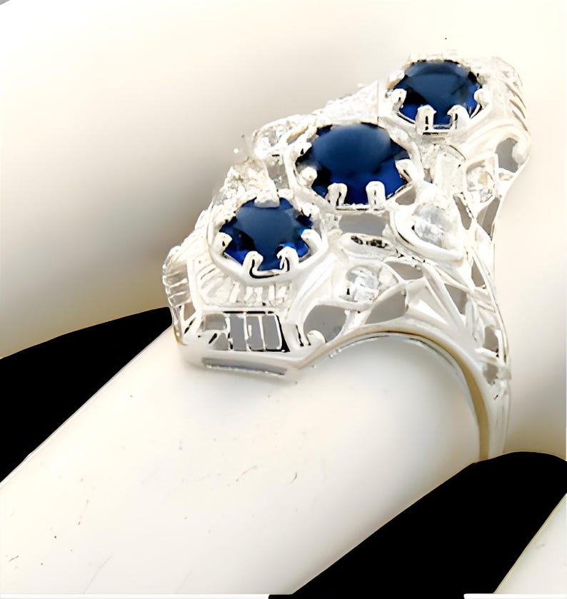 May include: A silver-toned ring featuring three round, deep blue faceted stones set in a row. The metal band displays an intricate, openwork filigree design with small, clear, colourless accent stones embedded throughout the decorative metal frame.