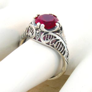 May include: A silver ring featuring a round, faceted, deep red gemstone set in a prong setting. The band is crafted with intricate, openwork metal filigree detailing that extends from the base of the stone setting down along the sides of the ring.