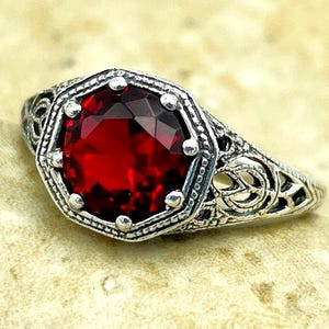 May include: A silver ring featuring a round, faceted deep red gemstone set within a hexagonal frame. The band and setting display intricate, openwork metal filigree detailing, creating a textured, decorative appearance around the central stone.
