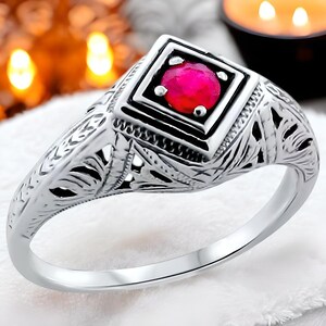 May include: A silver ring with an intricate filigree design and a square-shaped red gemstone in the center.