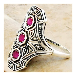 May include: An ornate silver ring featuring three oval, bright pink gemstones. The ring has an elongated design with intricate filigree and small, white pearl accents. The ring's band is smooth and polished.