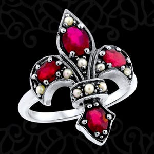 May include: A silver-toned ring featuring a fleur-de-lis design. The ring is adorned with oval-shaped red gemstones and small, round pearls. The ring has a polished finish and is set against a black background.
