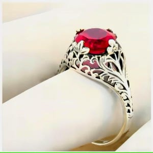 May include: A silver-toned ring featuring a large, round, faceted red gemstone set in a decorative, openwork metal band with intricate, leaf-like filigree patterns that extend from the central setting down along the sides of the ring shank.