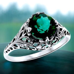 May include: A silver ring with a green gemstone set in a filigree design. The ring has a delicate, intricate design with a floral motif.
