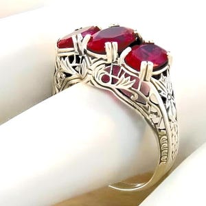 May include: A silver-toned ring featuring three oval-cut deep red gemstones set in a row. The band is crafted with intricate openwork metal detailing, showcasing delicate leaf-like patterns and filigree designs along the sides of the setting.