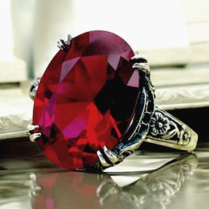 Puede incluir: Un anillo con una gran piedra preciosa de color rojo intenso y corte ovalado, sujeta por cuatro garras de plata. La banda de plata presenta un intrincado patrón floral en relieve en los laterales, con un acabado oscuro y oxidado.
