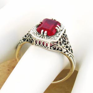 May include: A ring featuring a rectangular, deep red gemstone set in a silver-toned metal band. The setting displays intricate, openwork filigree detailing along the sides and around the central stone, which is secured by four small prongs.