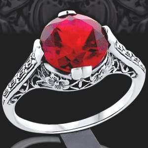 May include: A silver-toned ring featuring a large, round, faceted red gemstone set in a prong setting. The band is decorated with intricate, raised floral and scrollwork patterns that extend from the setting down the sides of the ring.