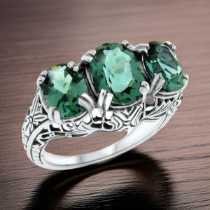 May include: A silver ring featuring three large, oval-cut green gemstones. The ring has an ornate, filigree design on the band and around the gemstones, with floral and scroll details. The gemstones are set in a prong setting.