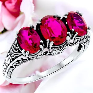 May include: A silver ring with three oval-shaped pink gemstones set in a filigree design.