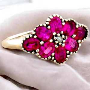 May include: A gold ring with a pink flower design made of ruby stones. The center of the flower has a pearl.