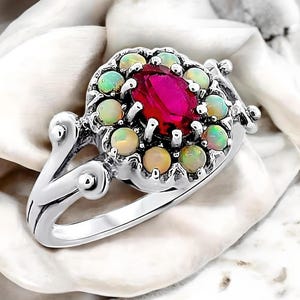 May include: A silver ring with a large red gemstone surrounded by a halo of smaller white opals. The ring has a delicate, ornate design.