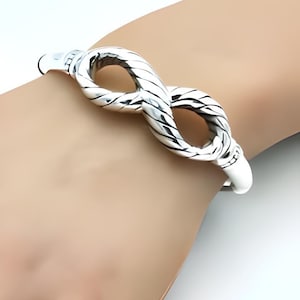 May include: A silver-colored bracelet with an infinity symbol design. The bracelet has a twisted rope-like texture and is worn on a wrist. The bracelet is made of a shiny metal.