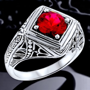 May include: A silver-toned ring featuring a square-cut, vibrant red gemstone set in the center. The band and setting are crafted with intricate, openwork filigree metal detailing that extends along the sides of the ring, creating a textured appearance.