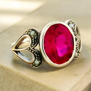 May include: A silver ring with a large, oval, bright pink gemstone as the centerpiece. The ring has ornate detailing on the sides, including heart-shaped designs and small, pearl-like accents. The ring is set on a neutral surface.