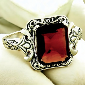 May include: A silver ring with an ornate design featuring a large, square, red gemstone.