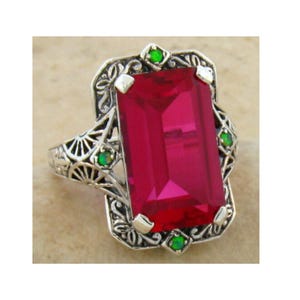 May include: A silver ring featuring a large, rectangular, ruby-red gemstone. The ring has an ornate, filigree design around the central stone, with small green gemstones accenting the corners and sides. The ring's band has a detailed, openwork pattern.
