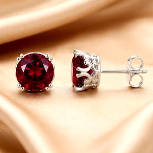 May include: A pair of stud earrings featuring round, deep red faceted stones set in silver-toned metal. The settings have an intricate, openwork scroll design along the sides, and the earrings include standard post backs for secure attachment.