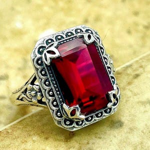 May include: A silver-toned ring featuring a large, rectangular, deep red faceted gemstone set in a decorative frame with scalloped edges and intricate floral metalwork along the sides of the band, showcasing a polished and detailed metal finish.