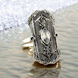 May include: A silver-toned ring with an elongated rectangular face. The ring features intricate etched patterns and a central, oval-shaped clear gemstone. The design includes floral and geometric elements, creating a vintage or Art Deco aesthetic.