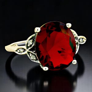 May include: A gold ring with a large, oval-shaped red gemstone surrounded by smaller, clear gemstones. The ring has a delicate, leaf-like design.