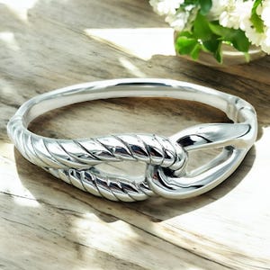 May include: A silver bangle bracelet with a knotted design. The knot is a rope-like design with a polished finish.