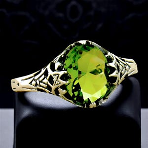 May include: A gold ring featuring an oval-cut, vibrant lime green gemstone set within an intricate, openwork metal band. The band displays a detailed, branching, or web-like pattern that extends from the central stone setting along the sides.