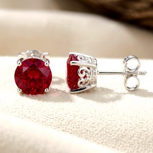 May include: A pair of stud earrings featuring round, deep red faceted stones set in polished silver metal. The settings have an intricate, openwork scroll design on the sides, and the earrings include standard silver post backs for secure wear.