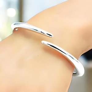 May include: A silver bangle bracelet with a curved, open design. The bracelet is made of a smooth, polished metal and has a simple, modern look.