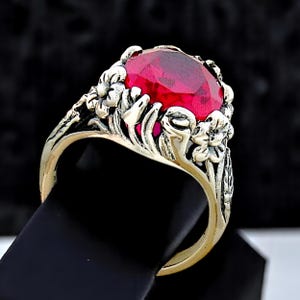 May include: A ring featuring a large, faceted, oval-shaped vibrant red gemstone set within a metallic band. The band is decorated with detailed, raised floral and leaf motifs that wrap around the sides of the central red stone setting.