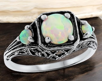 May include: A silver ring with an ornate filigree design. The ring features a large, round opal center stone, flanked by two smaller opals. The opals have a milky white and iridescent color. The ring is set on a dark wooden surface.