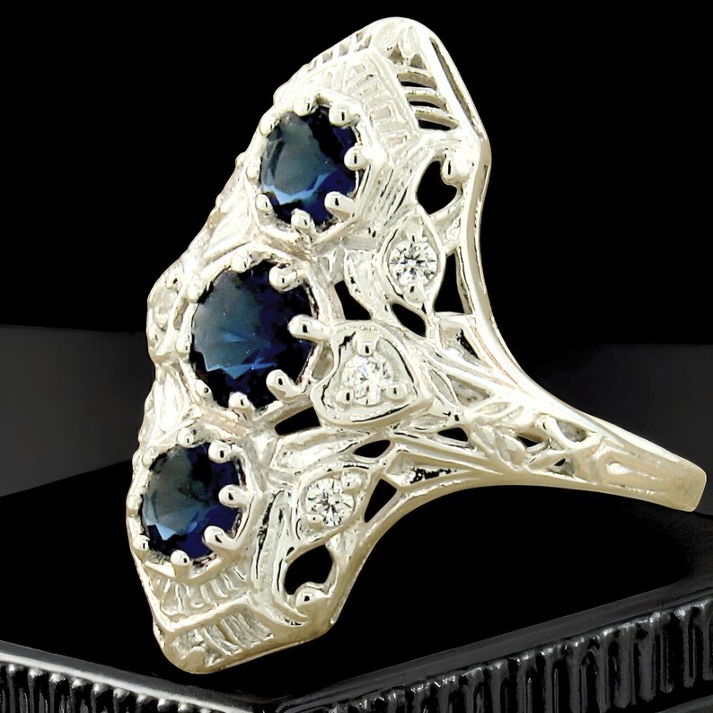 May include: A silver-toned ring featuring three round, deep blue faceted stones arranged vertically in a row. The metal band displays intricate openwork filigree detailing and is accented with small, clear, round stones set within the metal frame.