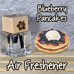 May include: A clear glass bottle with a wooden lid and a black reed diffuser stick. The bottle is labeled "Blueberry Pancakes" and "Air Freshener". The image also shows a stack of two pancakes with blueberry sauce and whipped cream.